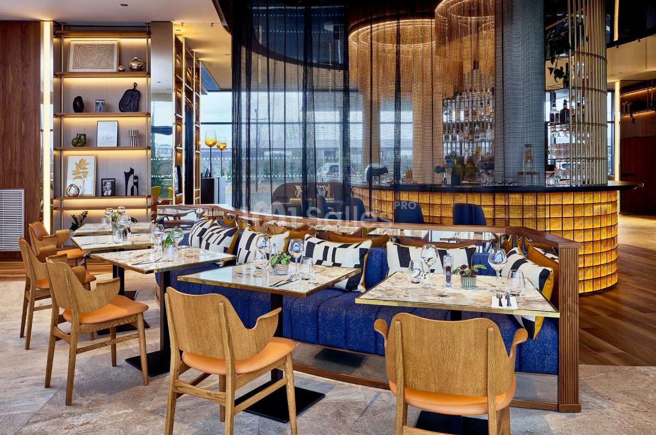 Salle de restaurant moderne avec tables dressées, banquettes bleues, chaises en bois et bar éclairé en arrière-plan.