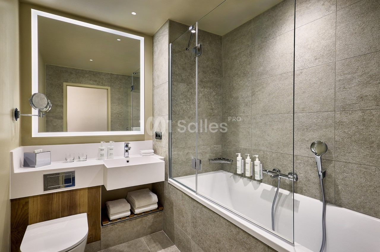 Salle de bain moderne avec baignoire, paroi vitrée, lavabo avec miroir éclairé et étagère contenant des serviettes.