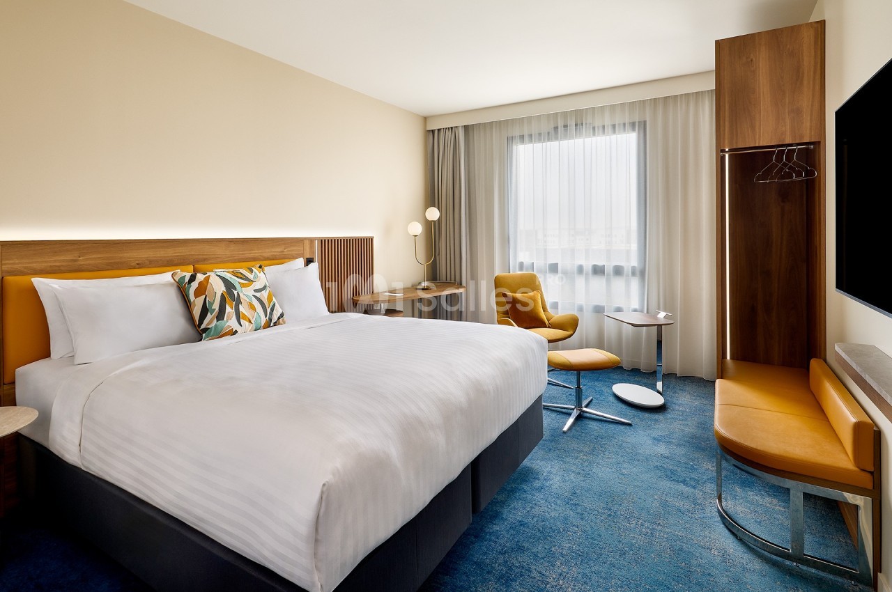 Chambre d'hôtel moderne avec lit double, fauteuil, bureau, téléviseur mural et grande fenêtre avec rideaux.