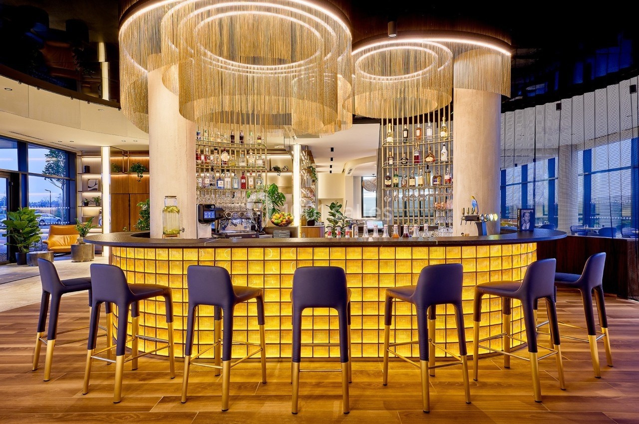 Bar moderne avec comptoir rétroéclairé, tabourets hauts bleus et décorations lumineuses suspendues.