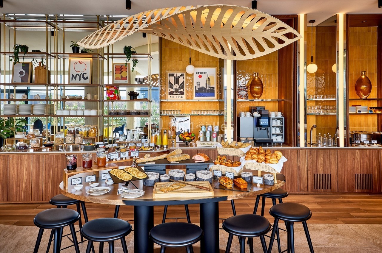 Buffet de petit-déjeuner avec viennoiseries, pains, confitures et boissons, dans un espace lumineux et moderne.