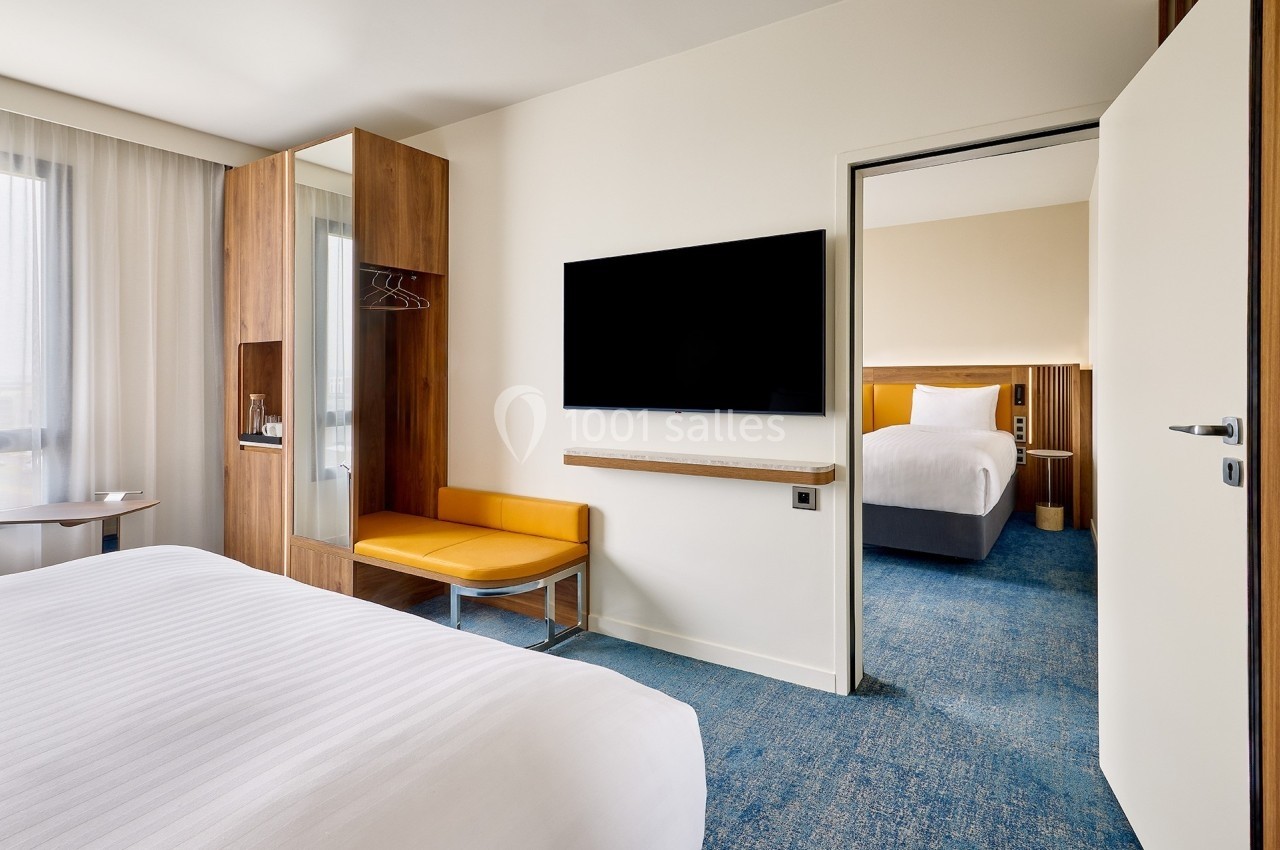 Chambre d'hôtel moderne avec lit, télévision murale, placard en bois et vue sur une seconde chambre adjacente.