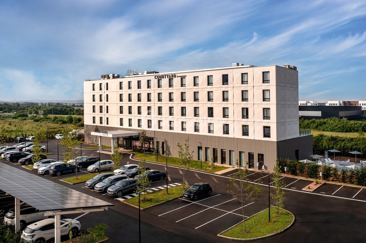 Façade d'un hôtel moderne avec parking attenant, entouré de verdure sous un ciel dégagé.