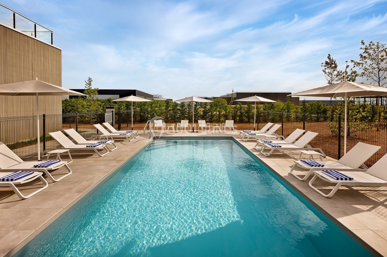 Piscine extérieure entourée de chaises longues et parasols, avec un ciel dégagé et un aménagement paysager en arrière-plan.