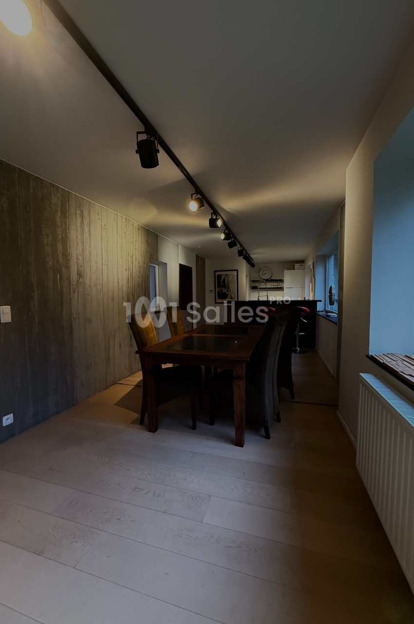 Salle à manger avec table en bois, chaises sombres, mur en bois à gauche et grande fenêtre à droite.