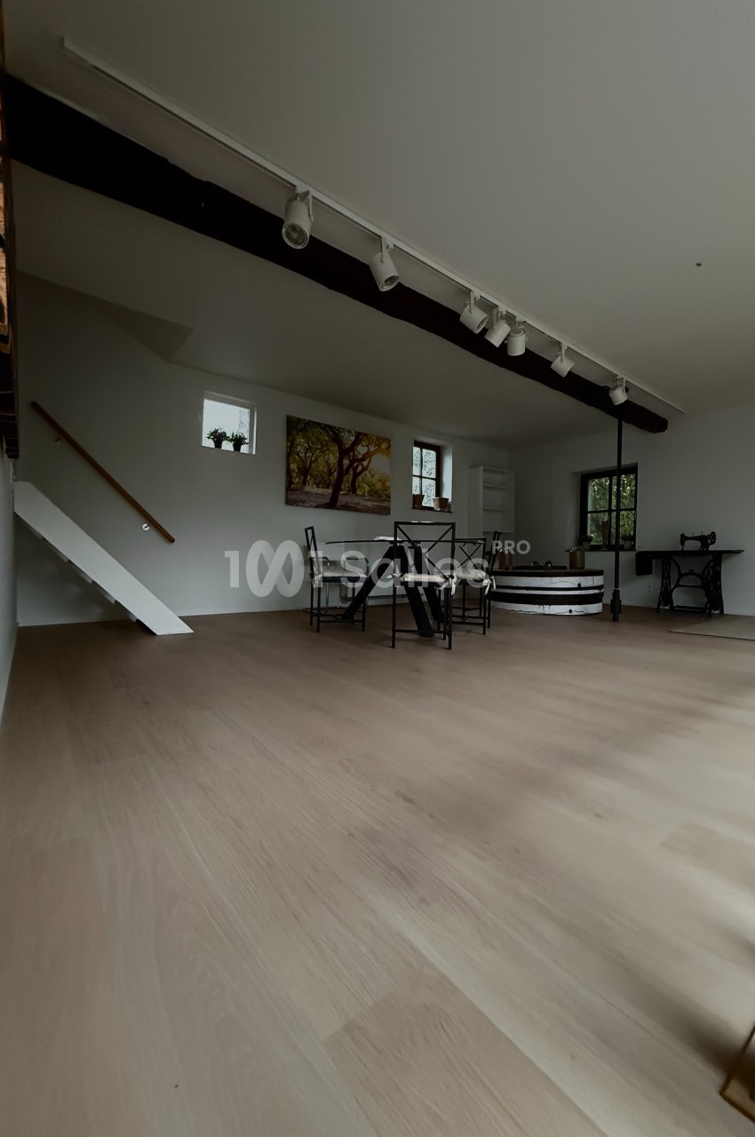 Salle lumineuse avec parquet clair, table et chaises au centre, escalier blanc et machine à coudre ancienne en arrière-plan.
