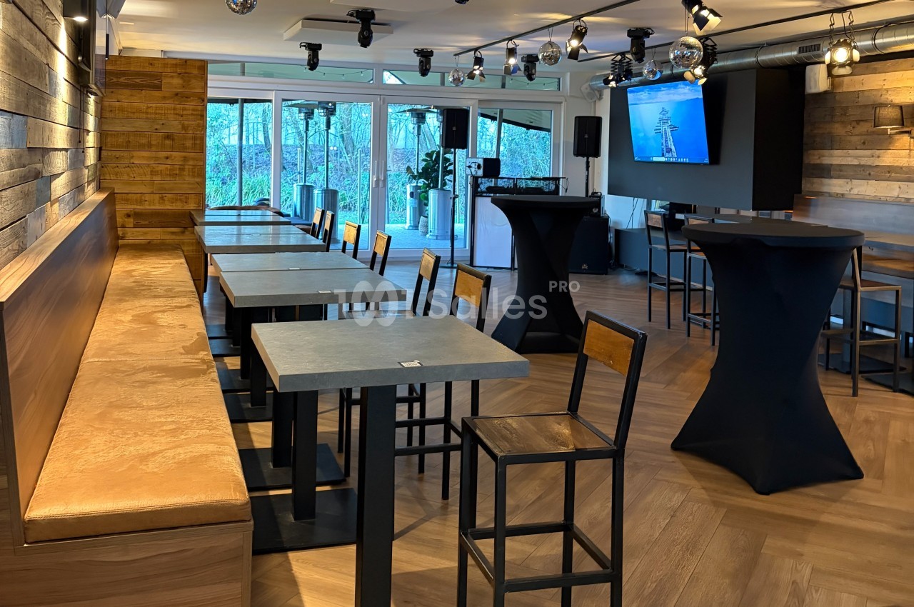 Salle lumineuse avec tables, chaises et mange-debout, décorée de bois et équipée d'un écran mural.