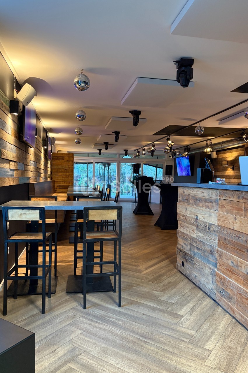 Intérieur d'un bar moderne avec mobilier en bois, parquet clair, tables hautes et écrans accrochés aux murs.
