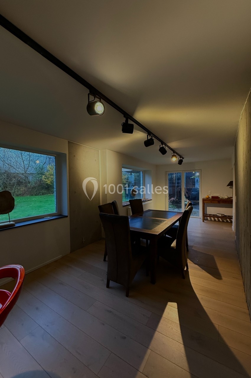 Salle à manger moderne avec table en bois, chaises sombres, éclairage sur rail et vue sur un jardin par de grandes fenêtres.