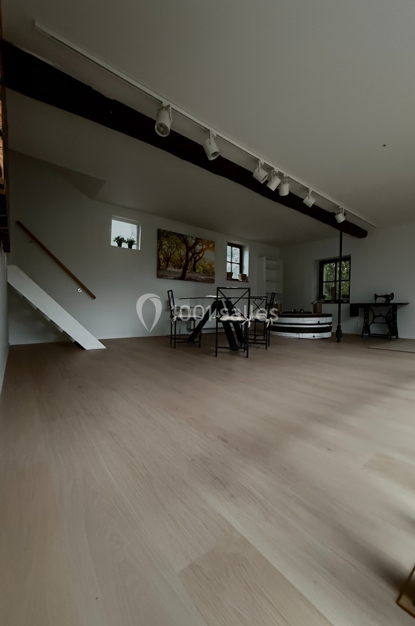 Salle lumineuse avec parquet clair, table et chaises au centre, escalier blanc, machine à coudre ancienne et tableau au mur.