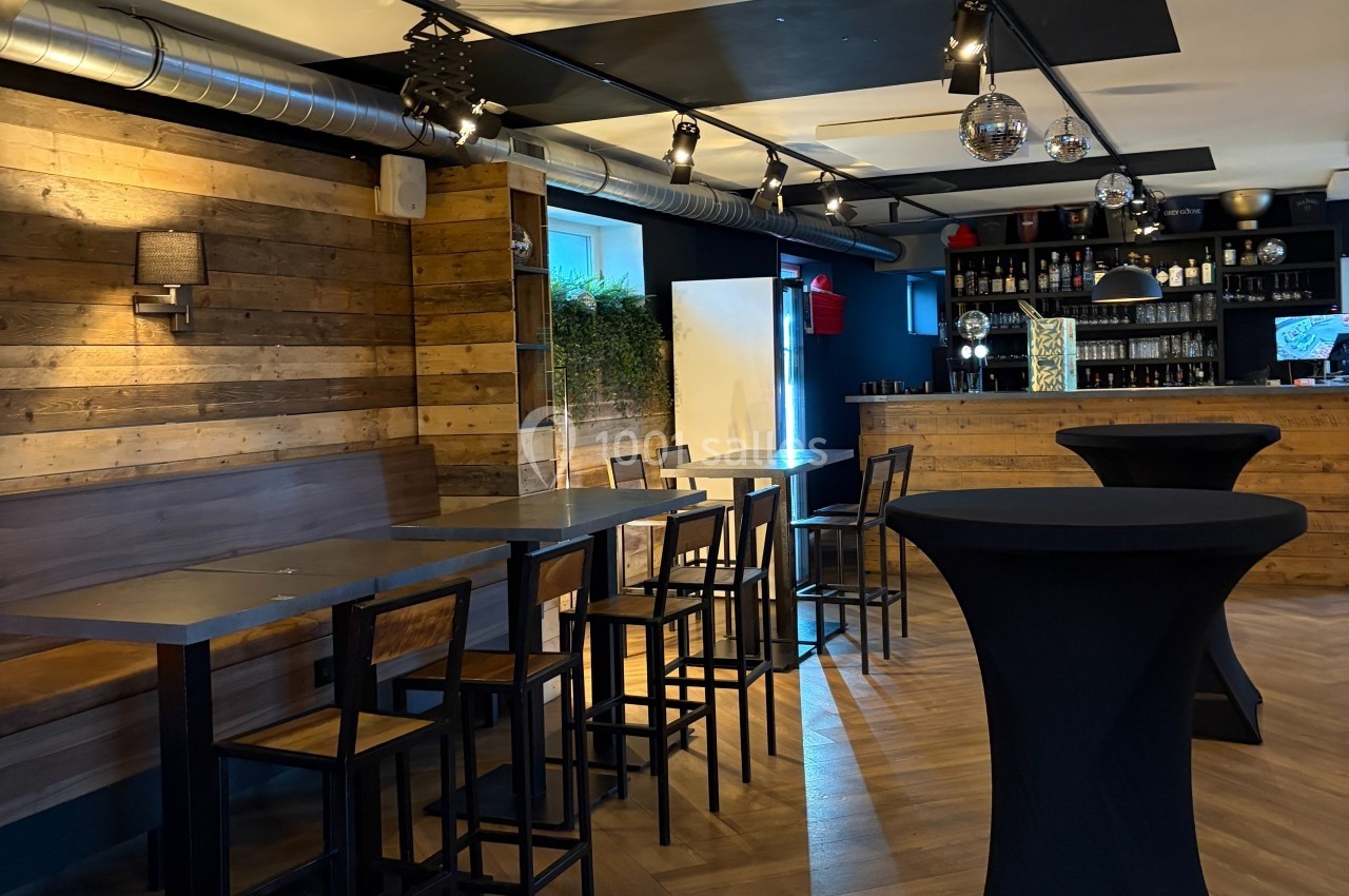 Salle moderne avec murs en bois, tables hautes, tabourets, bar en arrière-plan et éclairage tamisé.