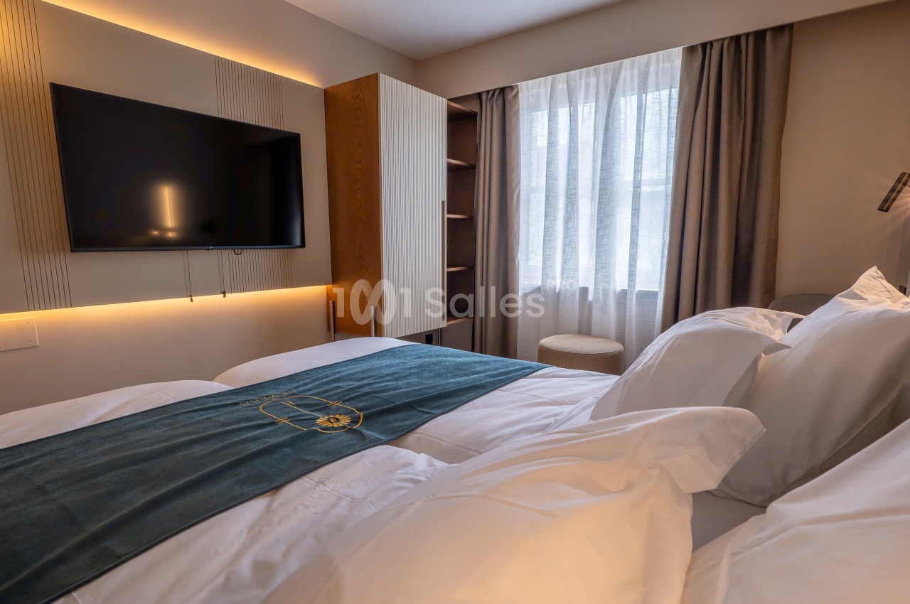 Chambre lumineuse avec lit double, télévision murale, rideaux épais et lumière naturelle entrant par une fenêtre.