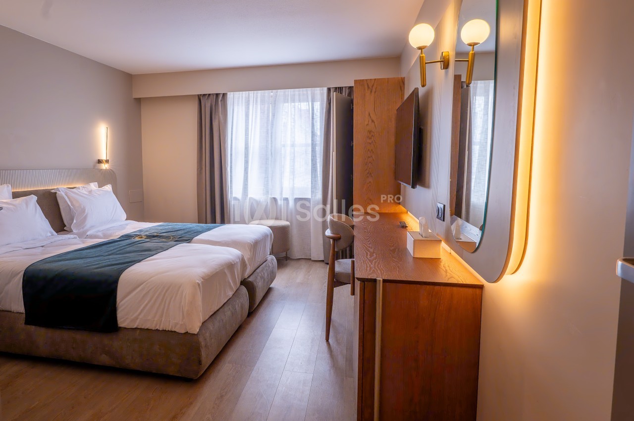 Chambre d'hôtel lumineuse avec lit double, bureau en bois, miroir éclairé et grande fenêtre avec rideaux.