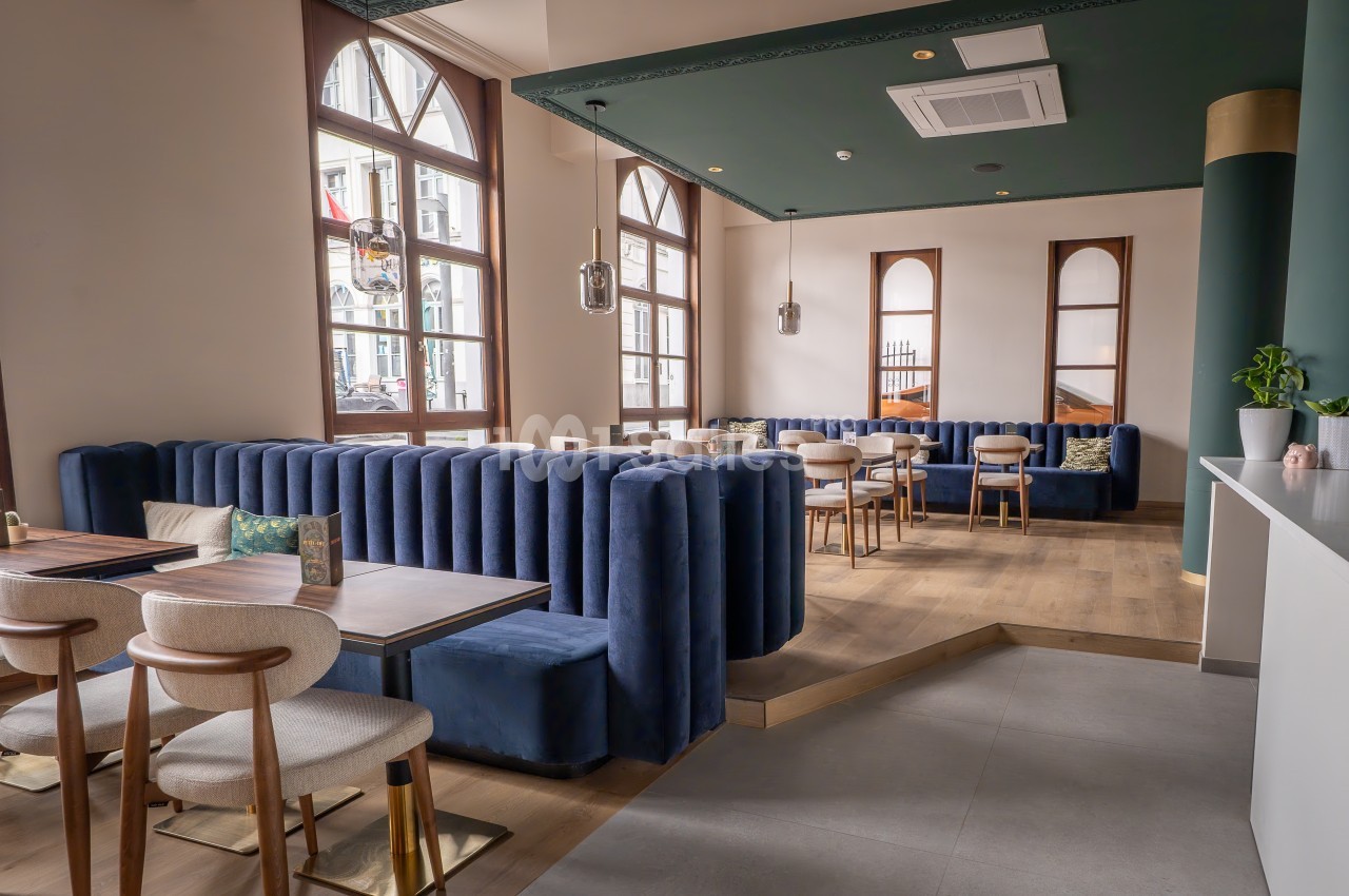 Salle de restaurant lumineuse avec banquettes en velours bleu, chaises en bois clair et grandes fenêtres en arc.