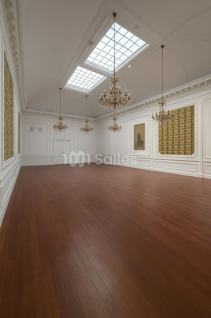 Salle lumineuse avec parquet en bois, murs blancs ornés de moulures, lustres suspendus et tableaux accrochés.