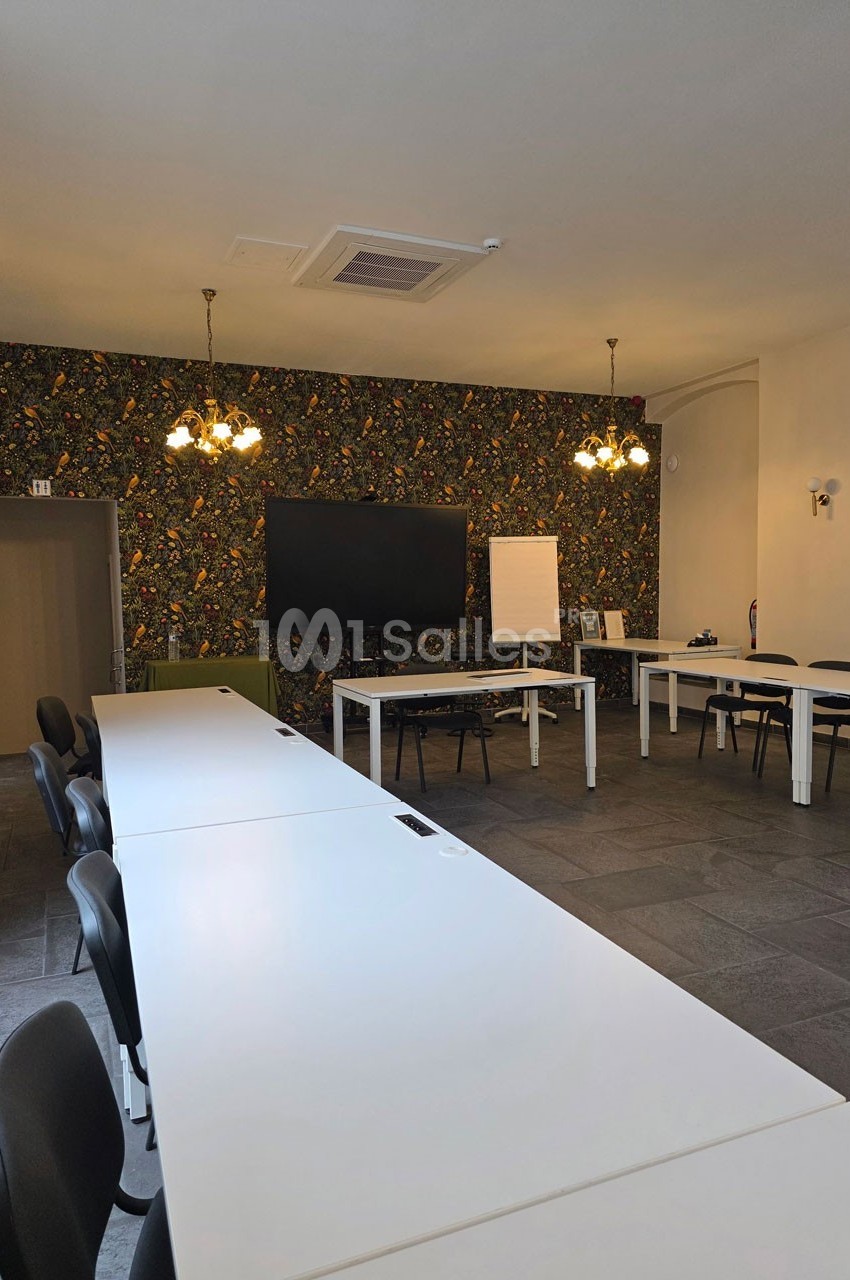Salle de réunion lumineuse avec de longues tables blanches, chaises noires, écran mural et tableau blanc.
