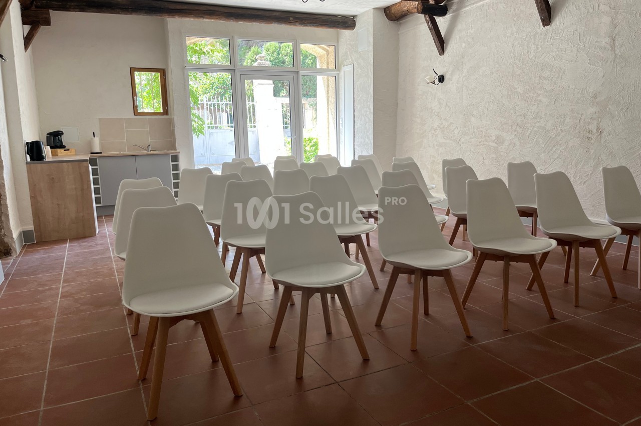 Salle lumineuse avec des chaises blanches alignées, sol en carrelage rouge et kitchenette en arrière-plan.