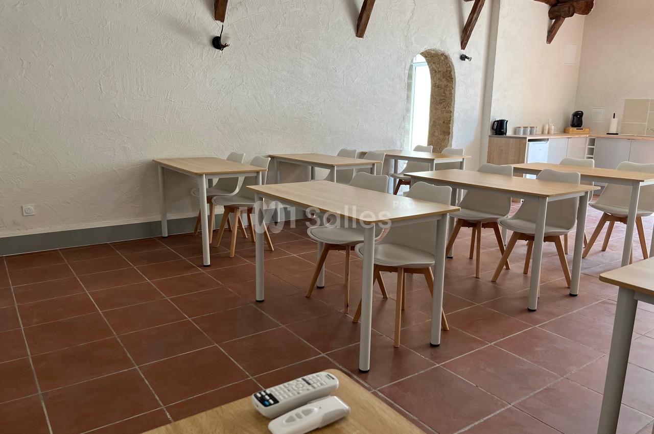 Salle lumineuse avec tables et chaises en bois clair, sol en carrelage rouge et coin avec cafetière en arrière-plan.