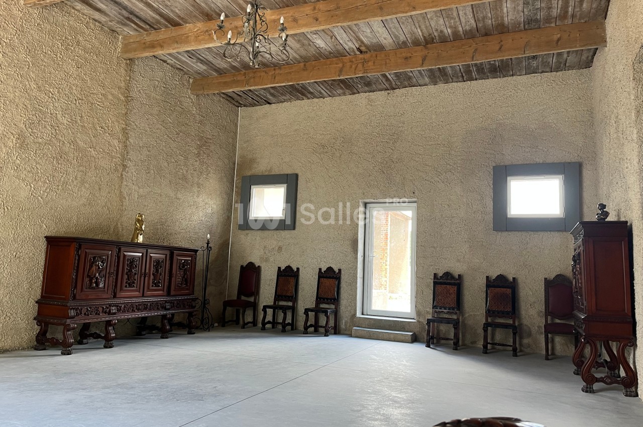 Salle spacieuse avec murs en pierre, plafond en poutres de bois, meubles anciens et chaises alignées contre le mur.