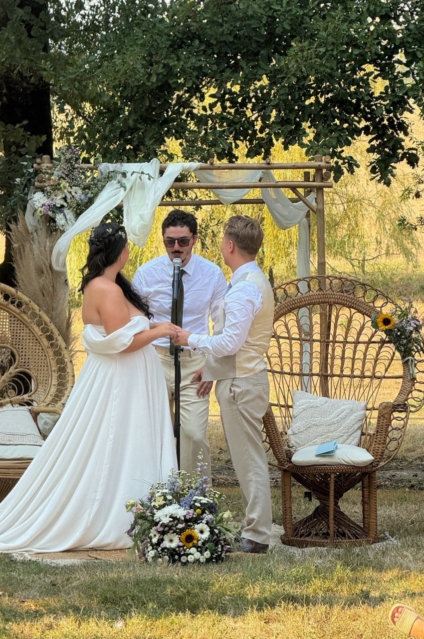 Un couple échange ses vœux devant un officiant, sous une arche décorée de fleurs, dans un cadre extérieur champêtre.