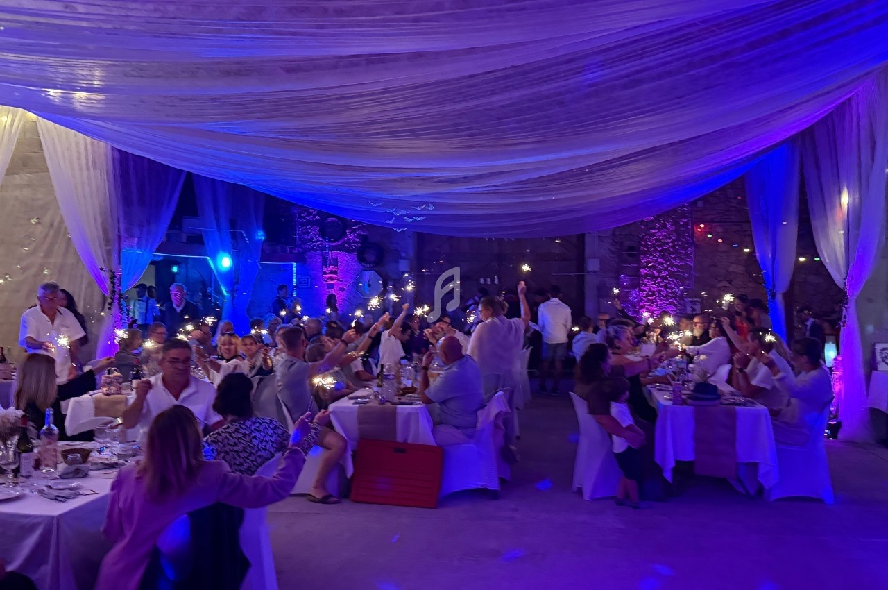 Des convives assis à des tables décorées, tenant des lumières, dans une salle éclairée par des tons violets et bleus.