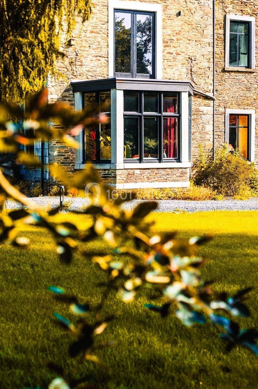 Façade en pierre d'une maison avec une grande baie vitrée, entourée de verdure et d'arbustes au premier plan.