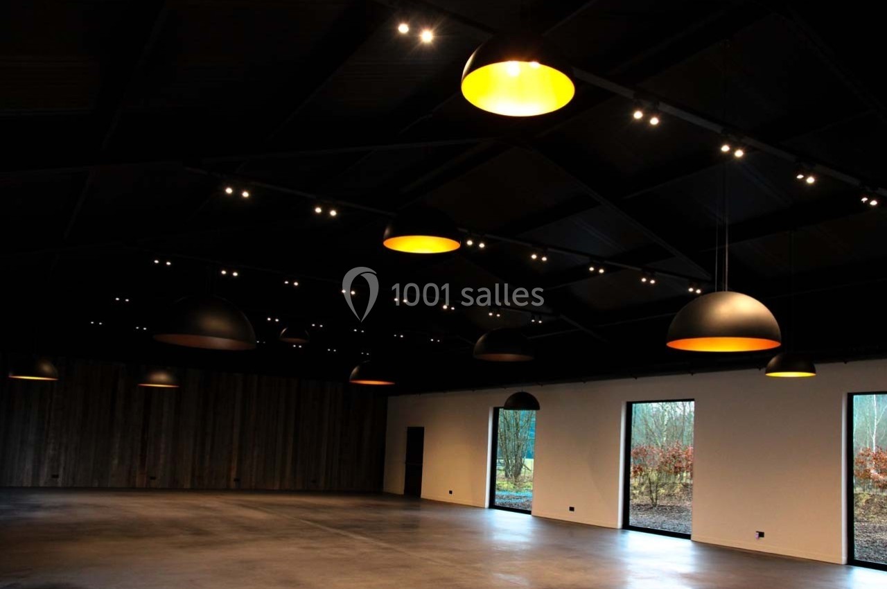Salle vide avec plafond sombre, éclairée par des suspensions lumineuses et des fenêtres donnant sur un extérieur arboré.