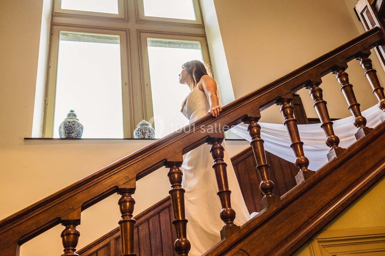Femme en robe blanche montant un escalier en bois, près d'une fenêtre avec deux vases décoratifs.