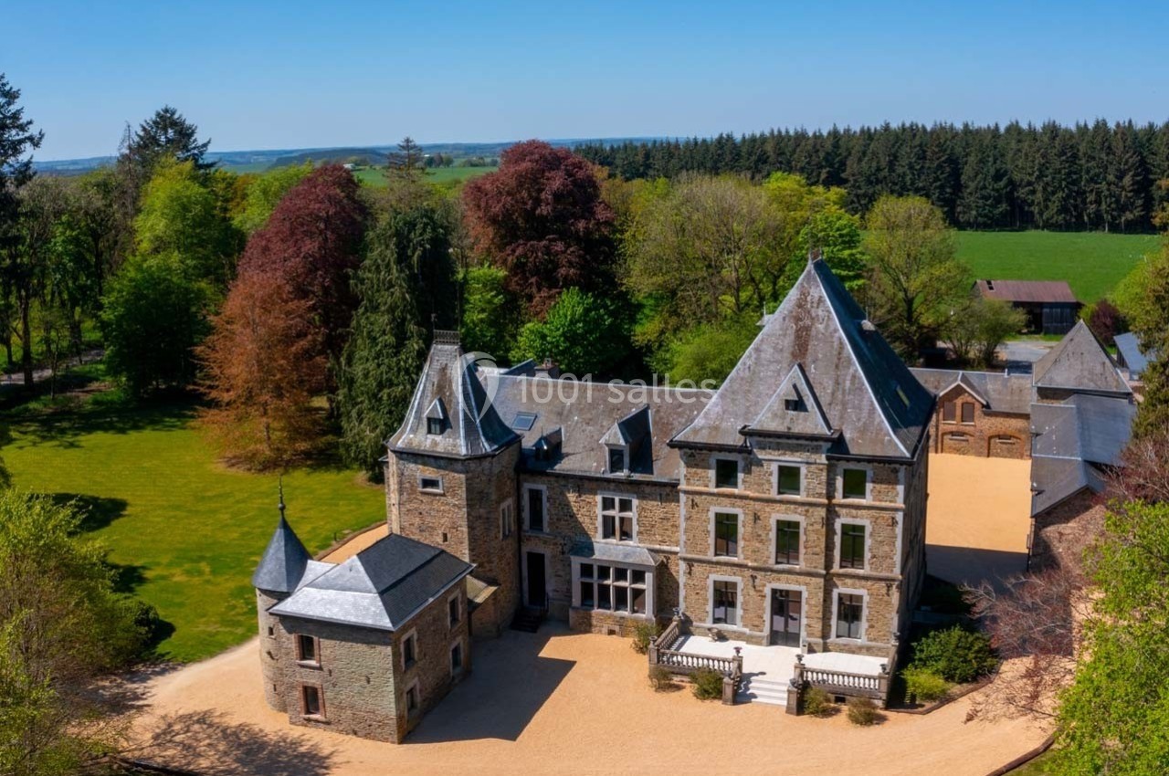 Vue aérienne d'un château en pierre entouré de pelouses, d'arbres et de forêts sous un ciel dégagé.