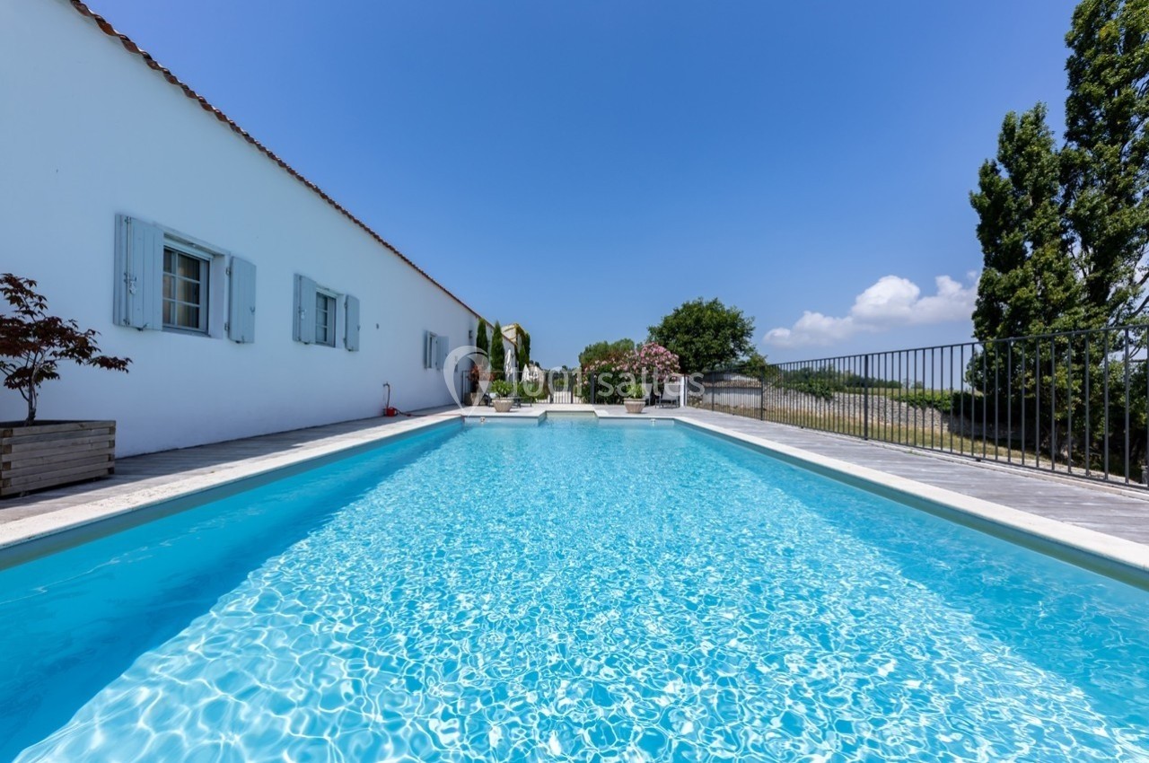 Piscine extérieure entourée d'une terrasse ensoleillée, avec une maison blanche et un ciel bleu en arrière-plan.