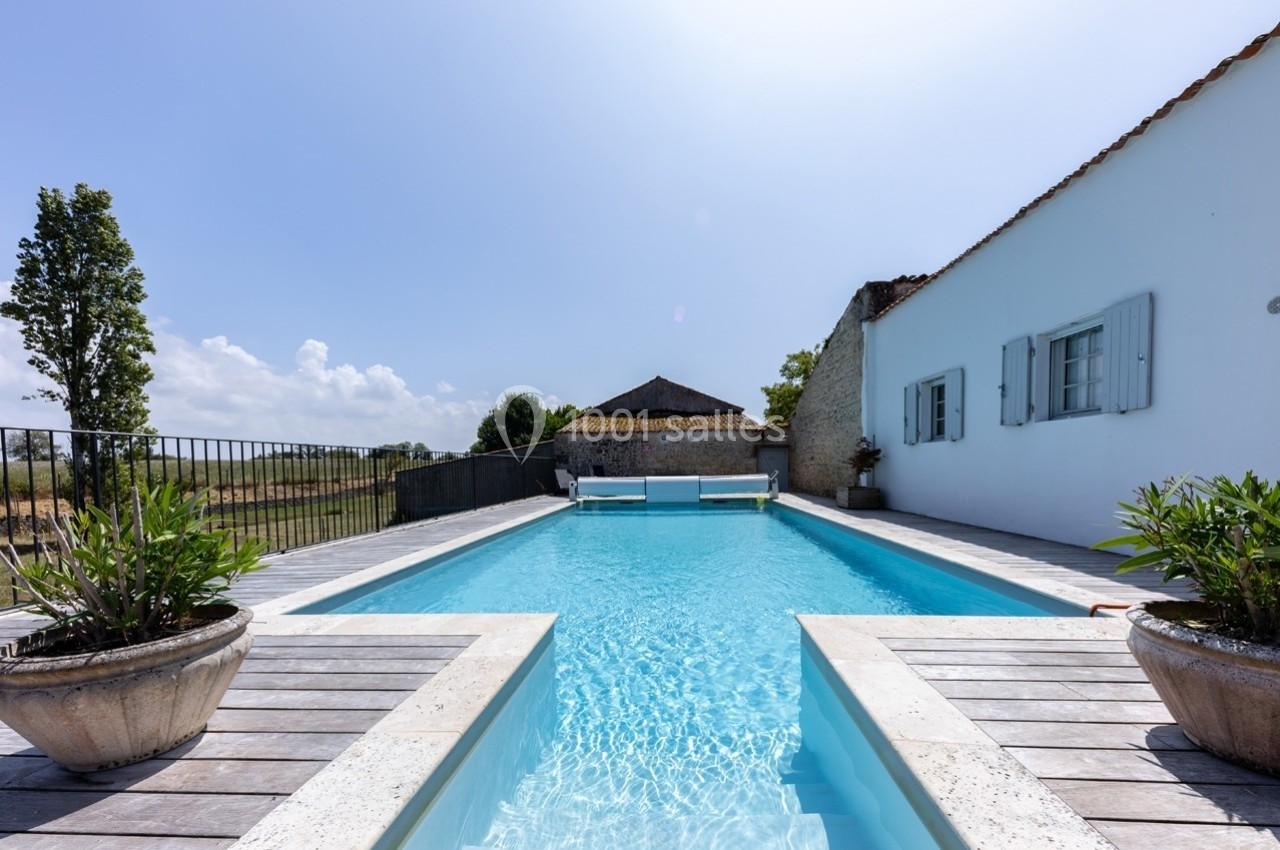 Piscine extérieure entourée de margelles en bois, avec une maison blanche et un paysage rural en arrière-plan.