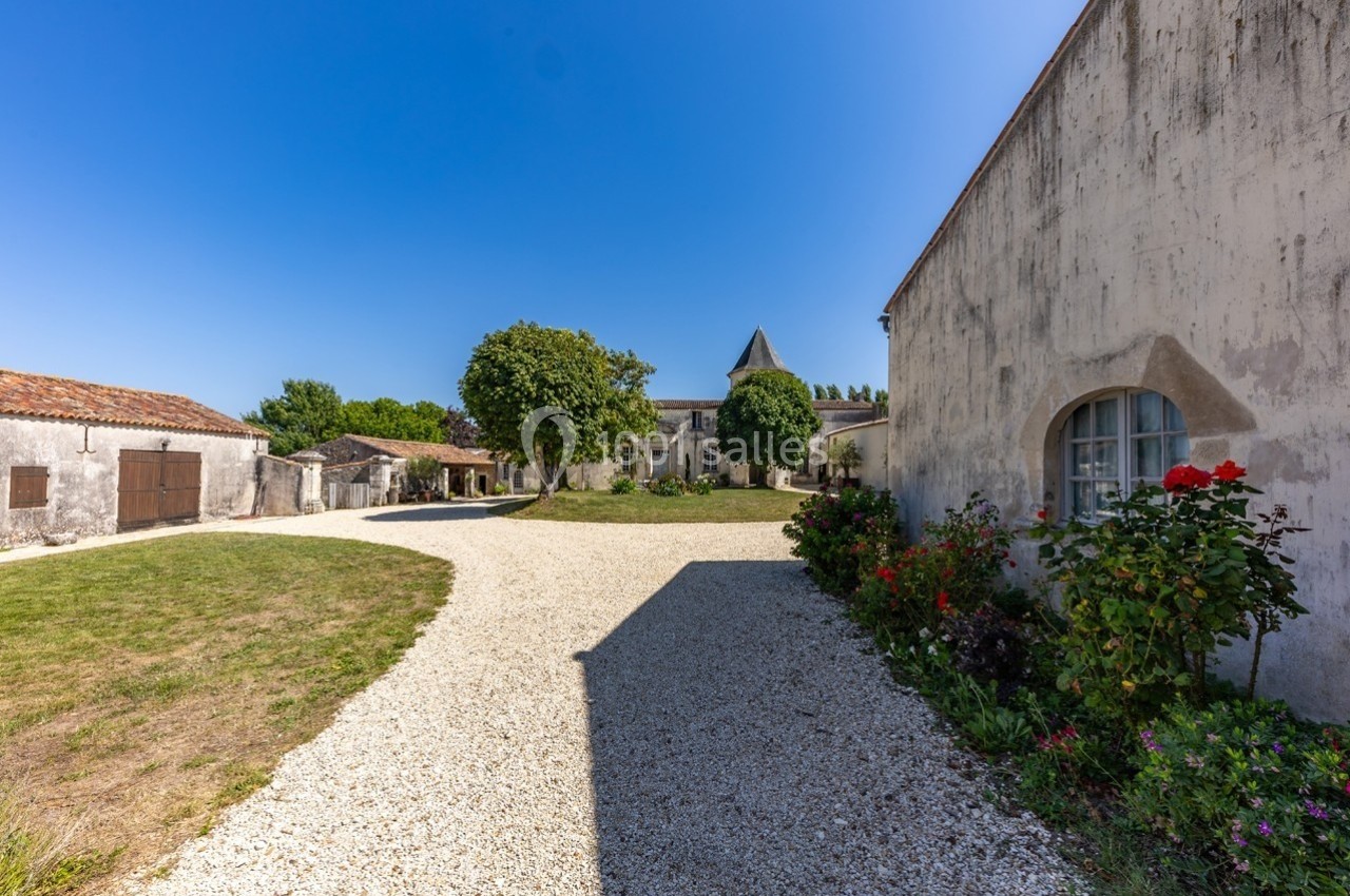 Cour en gravier bordée de bâtiments anciens, avec des arbres et des fleurs sous un ciel bleu dégagé.