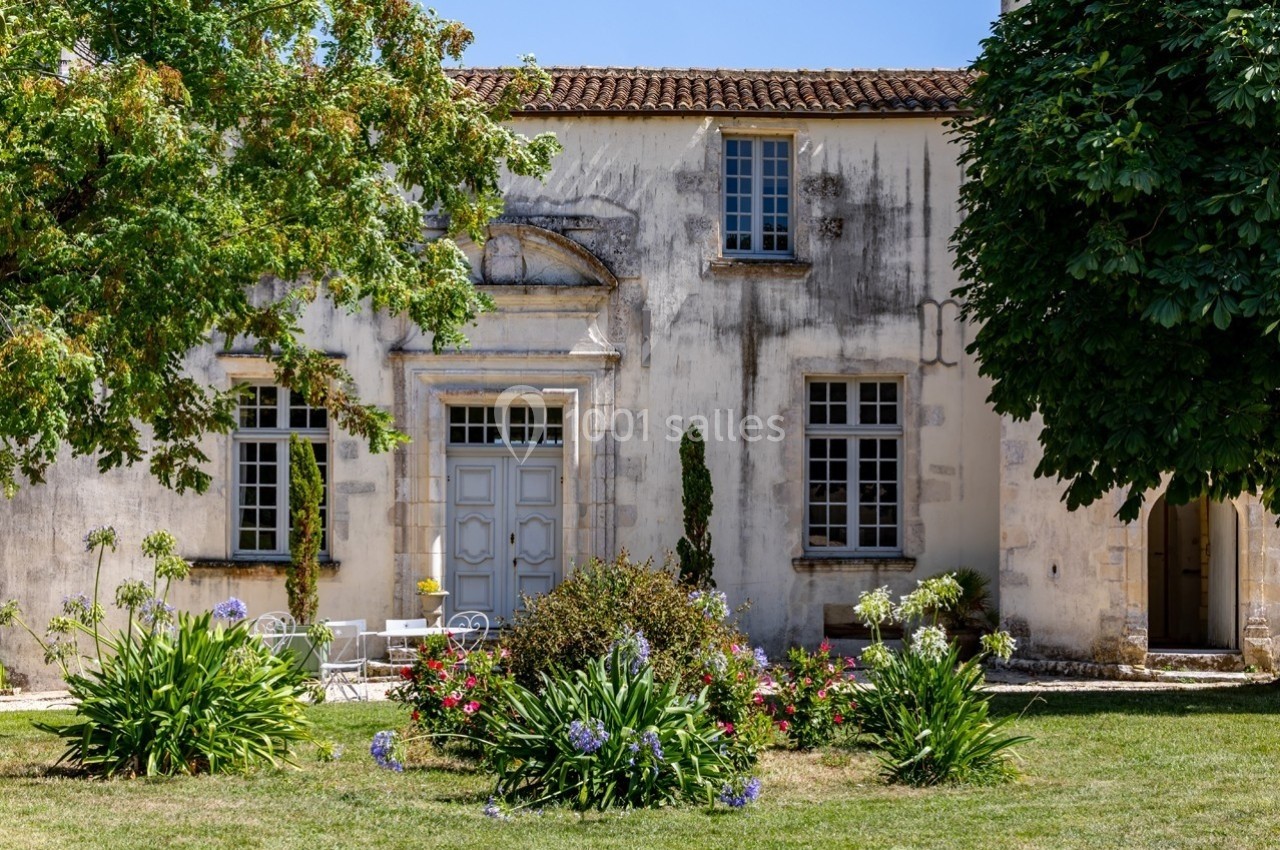 Façade d'une maison ancienne en pierre avec jardin fleuri, arbres et mobilier de jardin sous un ciel bleu.