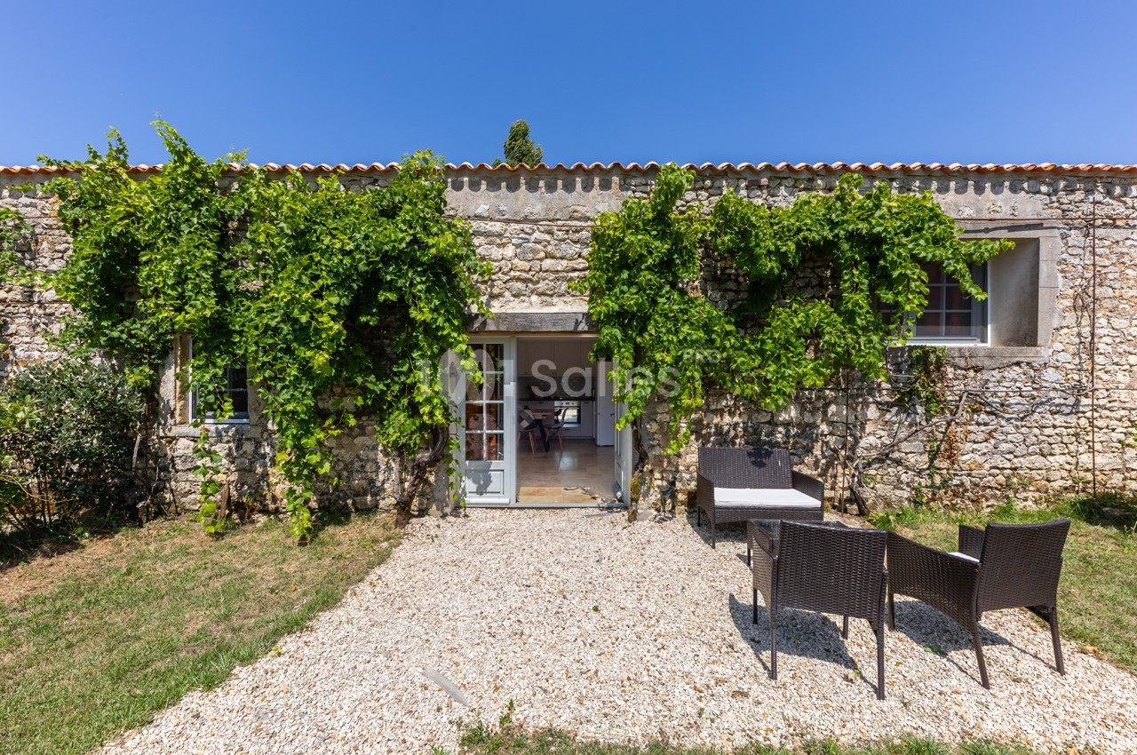 Façade en pierre d'une maison avec des plantes grimpantes, une porte ouverte et un salon de jardin sur gravier.