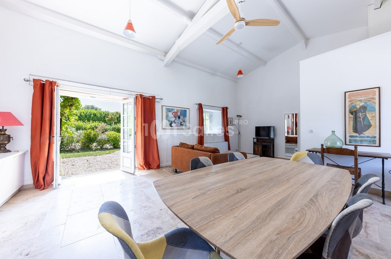 Salle à manger lumineuse avec table en bois, chaises colorées, canapé, et vue sur un jardin par une porte-fenêtre ouverte.
