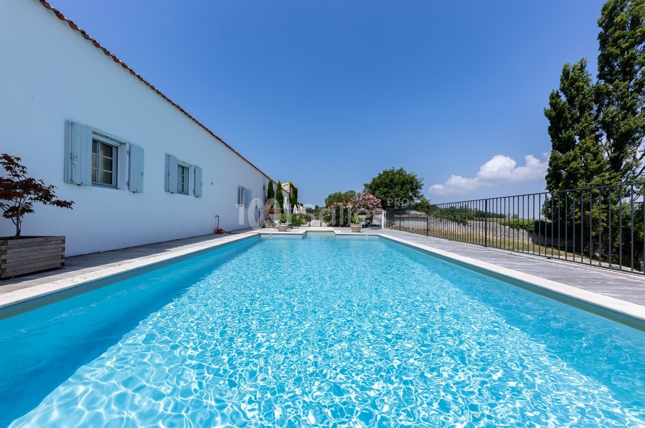 Piscine extérieure entourée d'une terrasse ensoleillée, bordée d'une maison blanche et d'arbres.