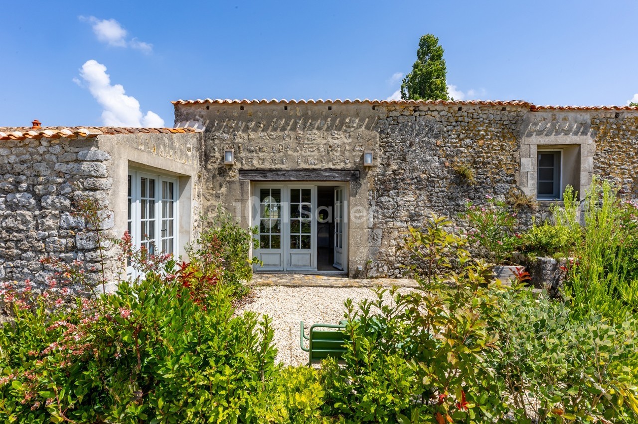 Façade en pierre d'une maison avec toit en tuiles, entourée de végétation et éclairée par un ciel bleu.