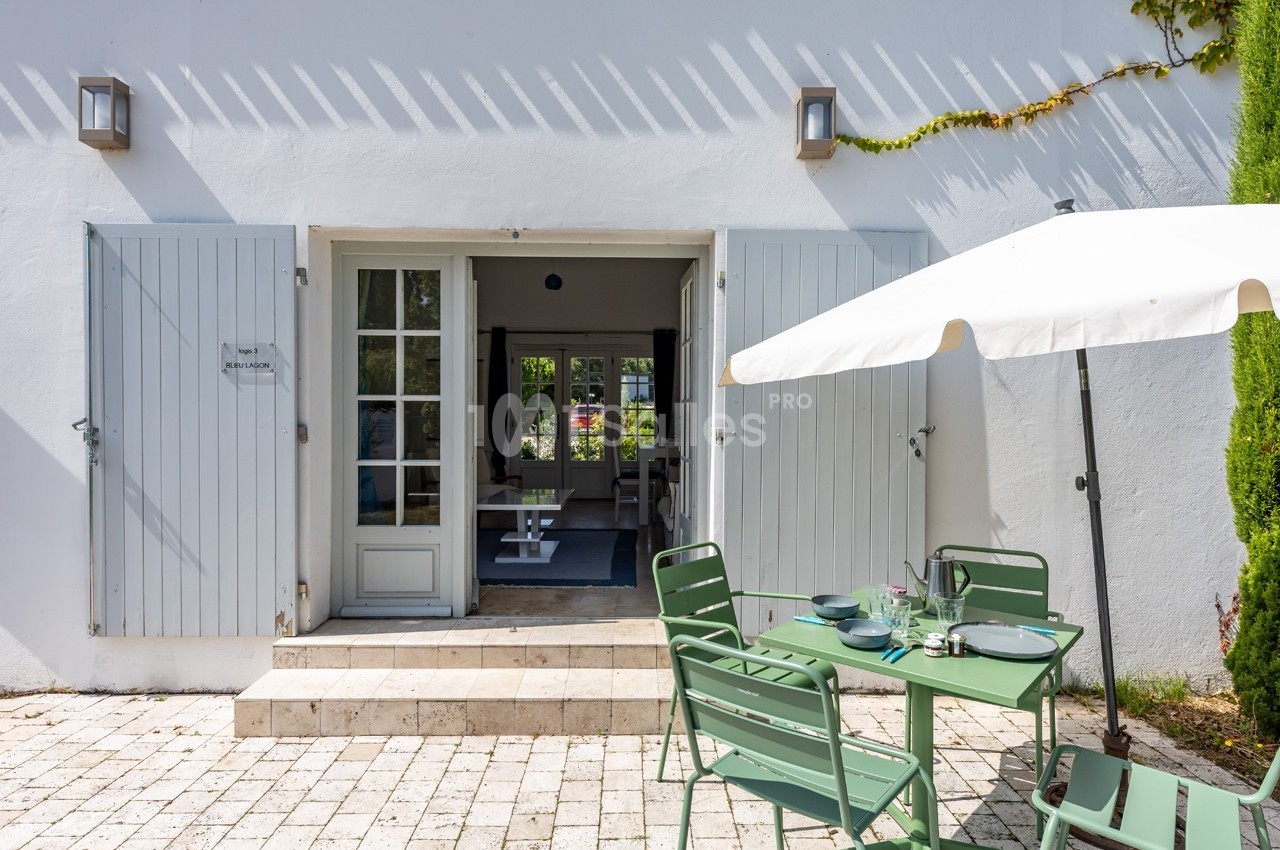 Terrasse ensoleillée avec table et chaises vertes, parasol blanc, et portes-fenêtres ouvertes sur un intérieur lumineux.