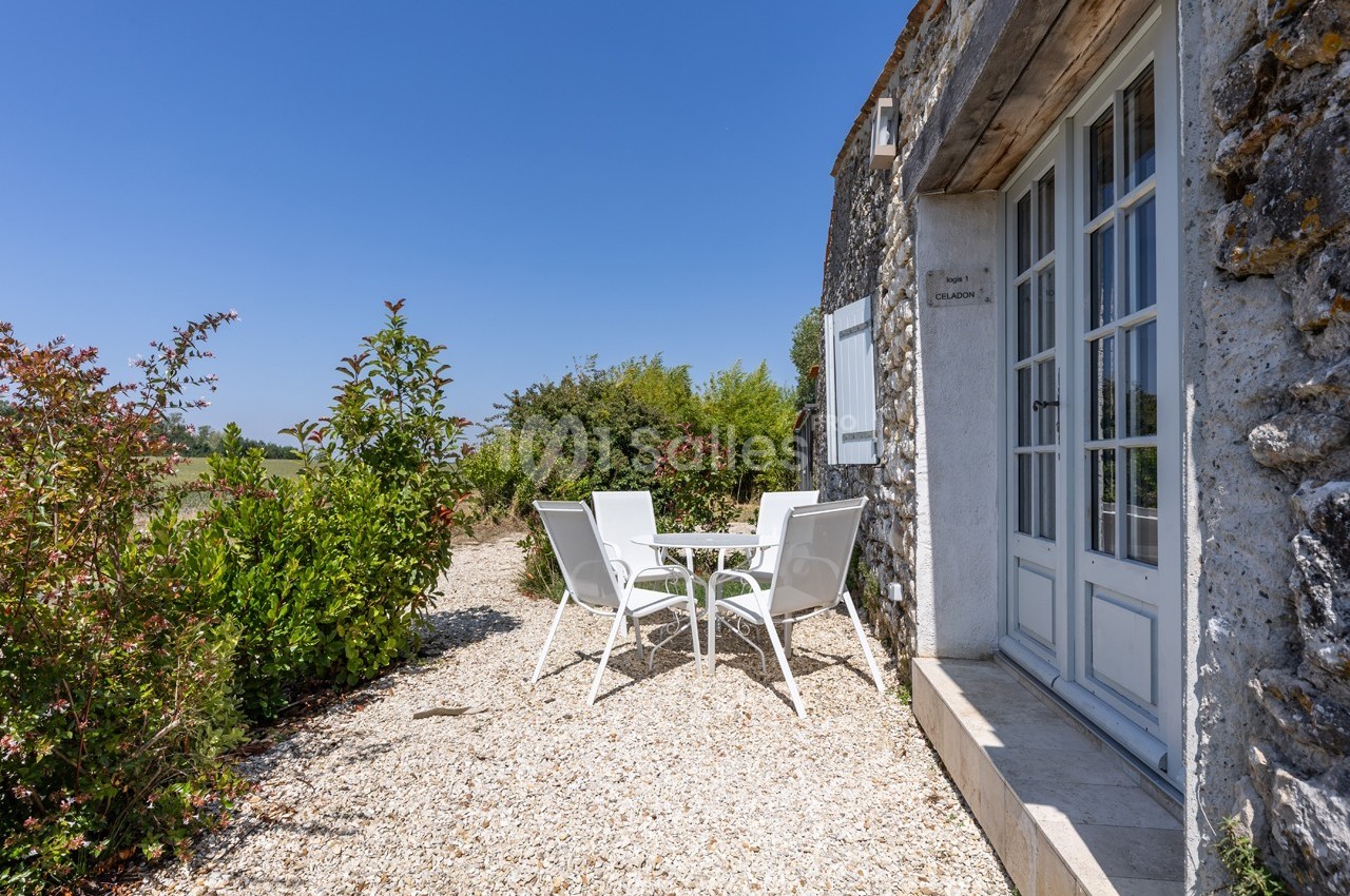 Table et chaises blanches sur une terrasse en gravier, entourées de végétation, près d'une maison en pierre.