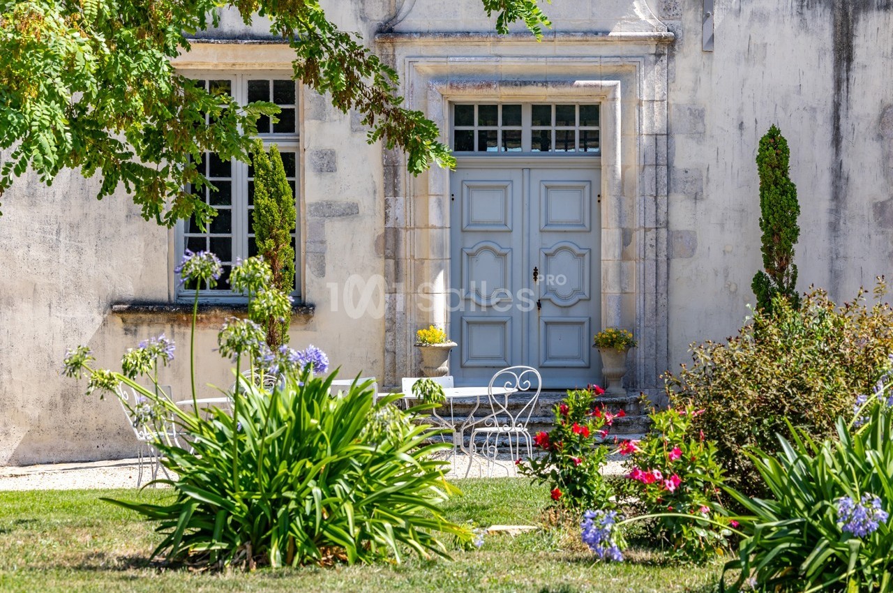 Façade d'une maison ancienne avec porte bleue, fenêtres à carreaux et jardin fleuri au premier plan.