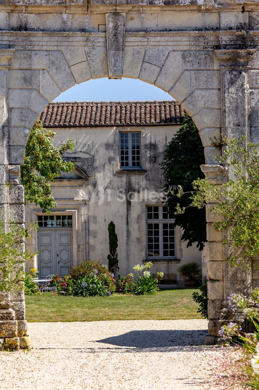 Vue d'une maison ancienne en pierre avec un jardin, encadrée par une arche en pierre.