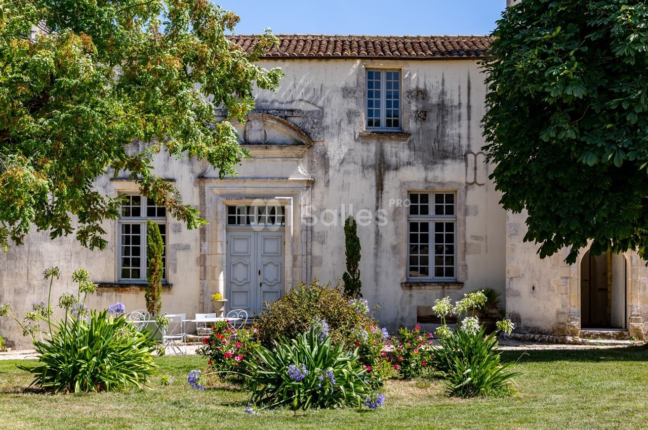 Façade d'une maison ancienne avec un jardin fleuri et des arbres sous un ciel bleu.