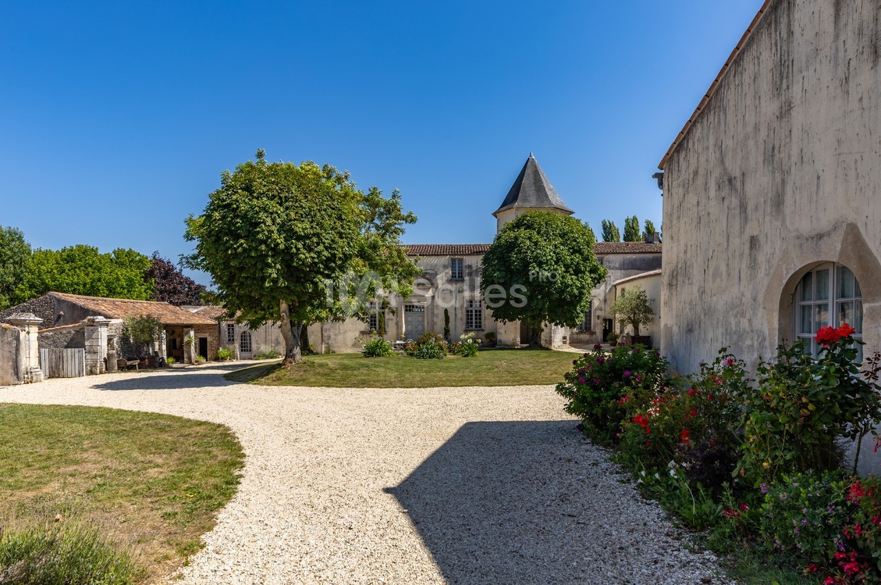 Cour d’un domaine rural avec bâtiments en pierre, arbres, pelouse et allée gravillonnée sous un ciel bleu.