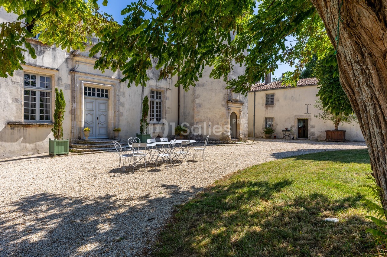 Cour d’un bâtiment ancien avec façade en pierre, table et chaises blanches au centre, entourée d’arbres et de verdure.