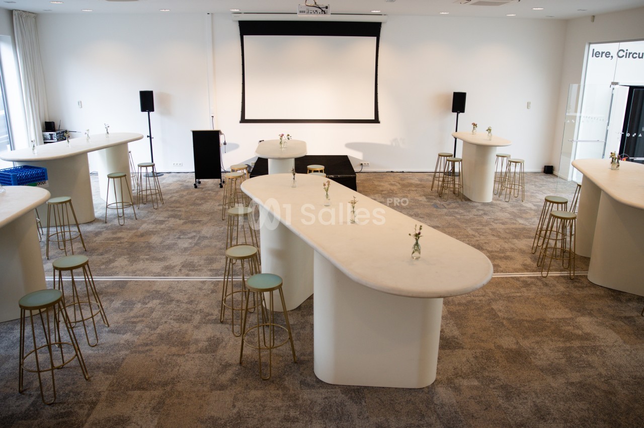 Salle lumineuse aménagée avec des tables hautes, des tabourets et un écran de projection en fond.