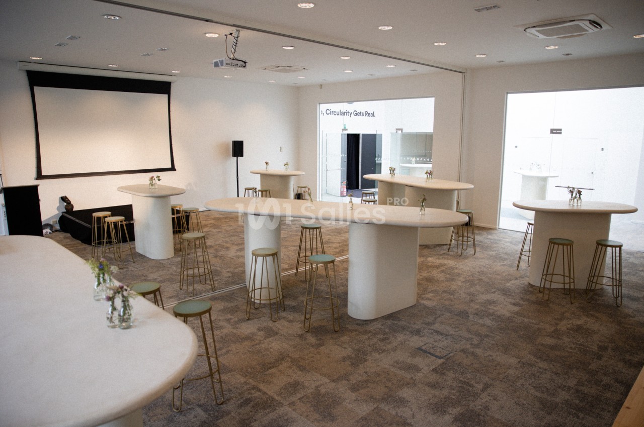 Salle lumineuse aménagée avec des tables hautes, des tabourets et un écran de projection en arrière-plan.
