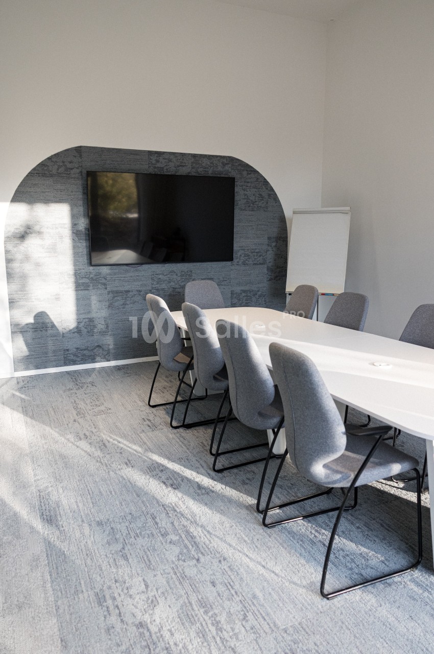 Salle de réunion lumineuse avec une table blanche, des chaises grises, un écran mural et un tableau de présentation.