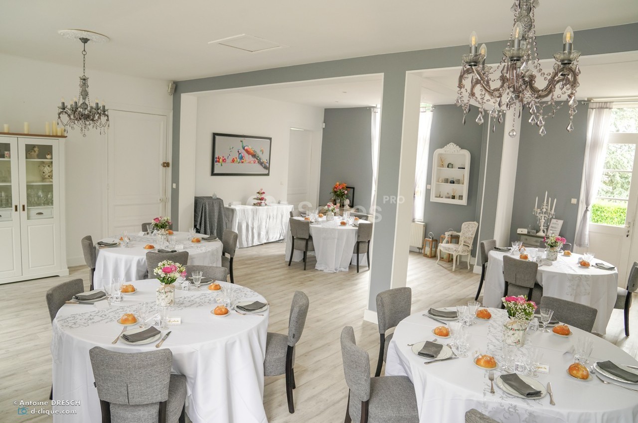 Salle lumineuse décorée pour un événement, avec des tables rondes dressées de nappes blanches et des chaises grises.
