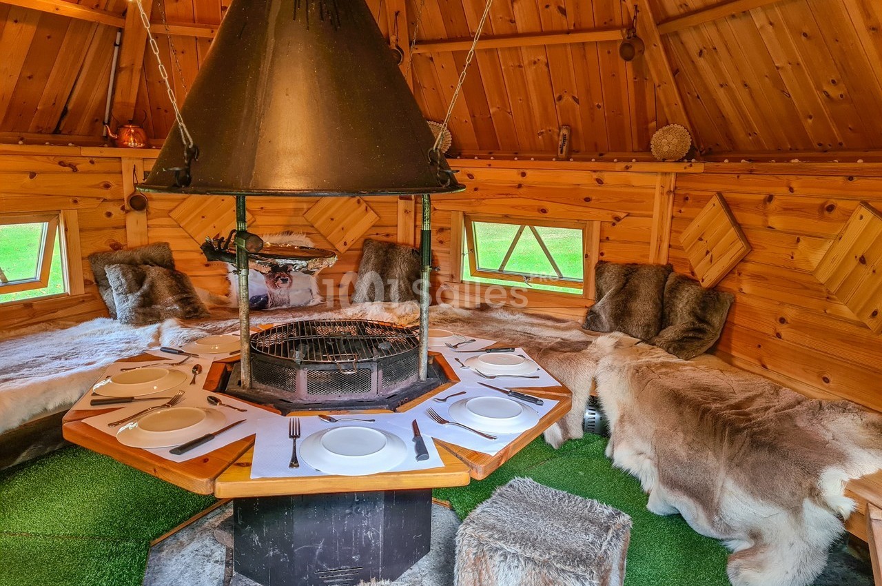 Intérieur d'un chalet en bois avec un barbecue central, des assises recouvertes de fourrures et une table dressée.