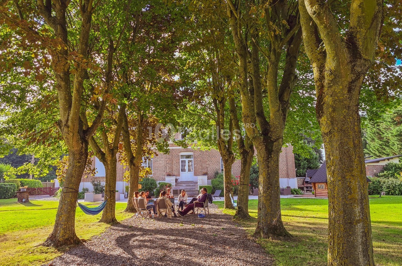 Groupe de personnes assises en cercle sous des arbres dans un jardin, avec une maison en briques en arrière-plan.